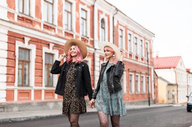 Gülümseyen, neşeli, pembe saçlı, şık yaz gençlik kıyafetleriyle saman şapkaları takan, el ele tutuşan ve açık havada gülümseyen ikizler. Moda elbiseli mutlu kız arkadaşlar yolda yürüyor.