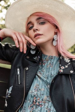 Pembe saçlı, şık hasır şapkalı, açık havada ahşap çitlerin yanında mavi elbiseli, siyah deri ceketli seksi bir kadın portresi. Çekici kız modeli kırsalda dinleniyor..