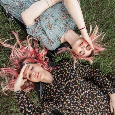 Pembe saçlı, şık yaz elbiseli ve yeşil çimenlerin arka planında seksi dudakları olan iki güzel genç kadın portresi. Sevimli kız arkadaşlar çimenlerde gözleri kapalı uzanır ve eğlenirler..