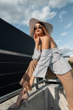 Modayı takip eden güneş gözlüklü, silindir şapkalı ve pantolonlu güzel bir kadın model şehirde ayakkabı giyiyor.