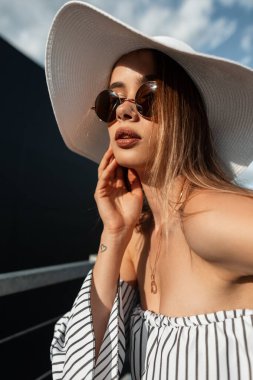 Şık çizgili bluzlu güneş gözlüklü, hasır şapkalı seksi dudaklı, çekici bir manken genç kadın şehirde güneşin altında poz veriyor. Şık yaz elbisesi içinde çekici bir kız güneşli bir günde dinlenmeyi sever..