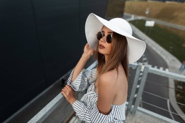 Havalı yaz kıyafetleri içinde şık bir genç kız. Şapkası, gözlüğü ve moda çizgili bluzu olan. Şehirde modern bir binanın yanında duruyor.