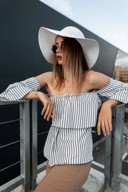 Moda mankeni, şık güneş gözlüklü, beyaz modaya uygun hasır şapkalı, zarif çizgili elbiseli, sokakta poz veren genç bir kadın. Açık havada poz veren havalı Avrupalı kız modeller. Vogue yaz kıyafeti giymiş güzel bayan..