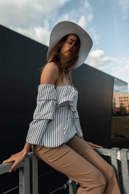 Güzel, genç ve çekici bir kadın, şık çizgili, şık bir şapkayla, bej pantolonlu, rüzgarlı bir yaz gününde modern binanın yakınında dinleniyor. Moda kıyafetli şık kız modeli dışarıda oturuyor..