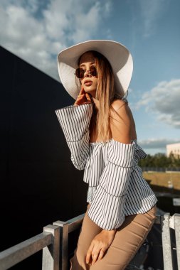 Havalı güneş gözlüklü, yaz şapkalı, çizgili bluzlu ve pantolonlu güzel hippi kadın güneşli bir günde, gökyüzü ve güneş ışığıyla şehirde oturuyor.