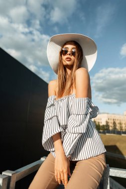 Moda mankeni, şık güneş gözlüklü, modaya uygun hasır şapkalı genç bir kadın açık havada güneşli bir yaz gününde dinleniyor. Kıyafetli güzel kız demir parmaklıkların yanında güneşlenmeyi sever.