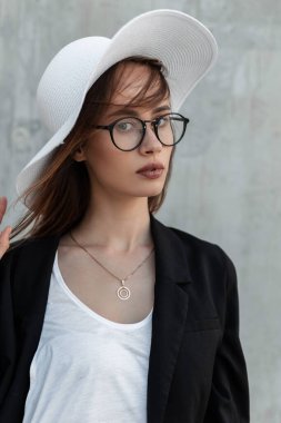 Güzel bir portre. Güzel dudakları, modaya uygun gözlükleriyle şık siyah-beyaz giysileri ve klasik gri arka planda yaz modası şapkası olan güzel bir genç kız..