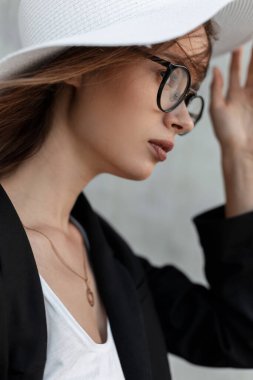Moda portresi, güzel dudakları olan, şık gözlüklü siyah elbiseli, saman beyazı zarif yaz şapkalı, gri duvarın yanındaki sokakta. Profil fotoğrafı.