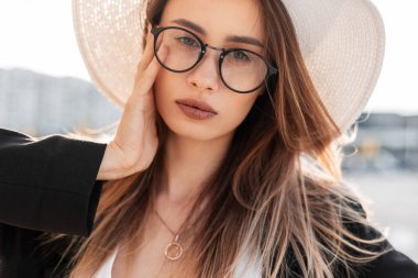 Açık havada güneşli bir günde ceketli, ceketli gözlüklü, şapkalı, güzel, seksi dudakları ve temiz ciltli çekici genç bir kadın portresi. Güzel şehir kızı yürümeyi ve gün ışığını sever..