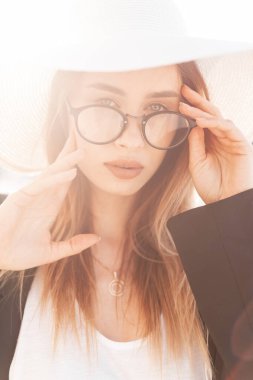 Plaj şapkalı, gözlüklü, siyah ceketli, gün batımında beyaz tişörtlü güzel bir kadın. Güneşli bir günde yaz kıyafetleri içinde oldukça şık bir kız.