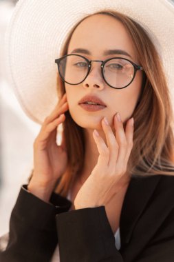 Gün batımında siyah ceketli, saman beyazı şapkalı, moda gözlüklü güzel bir genç kadın portresi. Güneşli bir günde sokakta zarif bir kız. Kadınlar için moda yaz kıyafetleri.