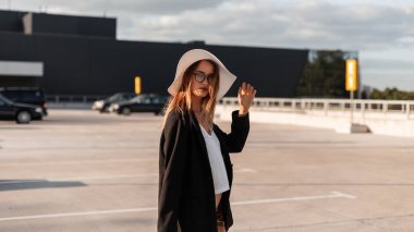 Moda modası geçmiş siyah ceketli genç bayan ve yazın şık bluzlu güzel şapkalı kadın güneşli bir günde açık havada poz veriyor. Otoparkta iş kıyafetleri içinde modaya uygun bir kız modeli. Zarif görünüm..