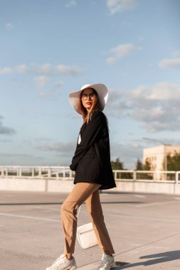 Moda gözlüklü, modaya uygun kıyafetler ve hasır şapkalı modern zarif genç kadın şehrin arka planında asfalt üzerinde yürüyor..