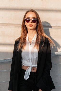 Güneşli portre oldukça seksi stil sahibi güneş gözlüklü şık siyah ceketli, açık havada duvarın yanında zarif beyaz gömlekli bir kadın. Günbatımında gündelik kıyafetlerle çekici bir manken kız..