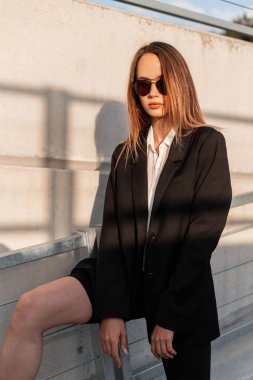 Şık genç, şık siyah elbiseli, şık güneş gözlüklü Amerikalı genç kadın açık havada yaz güneşinin gölgesinde duruyor. Modern model günbatımında sokakta yürümekten hoşlanıyor.. 