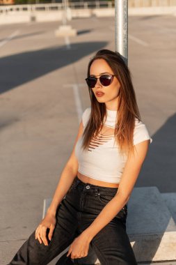 Klasik güneş gözlüklü, havalı siyah beysbol şapkalı, yaz kıyafetleriyle genç ve güzel bir hippi kadın güneşli bir günde asfaltta dinleniyor. Kentsel Amerikalı kız model..