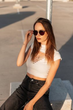Beyzbol şapkalı şık genç hippi kız güneşli bir günde moda güneş gözlüğünü düzeltiyor. Günbatımında şehrin otoparkında poz veren modaya uygun gençlik kıyafetleriyle güzel bir şehir kadını modeli..
