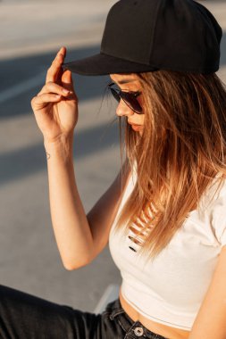 Profil portresi, siyah güneş gözlüklü, beyaz seksi bluzlu, şehirde şık şapkalı havalı şehir modasına uygun havalı bir kadın. Güneşli bir günde güzel şehir kızı otoparkta poz veriyor..