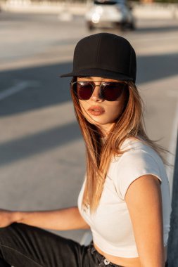 Güneşli bir portre. Güneş gözlüklü, şık siyah şapkalı, beyaz yırtık tişörtlü güzel bir kadın. Parlak yaz günbatımında otoparkta. Moda mankeni kız şehirde güneşte dinleniyor..