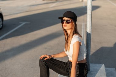 Modellik yapan siyah güneş gözlüklü, havalı siyah şapkalı yaz kıyafetleriyle genç bir kadın park yerinde dinleniyor ve güneşli bir günün tadını çıkarıyor. Amerikan modern görünümü.