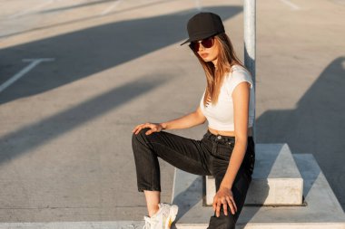 Moda siyah şapkalı, seksi beyaz bluzlu, modaya uygun kot pantolonlu ve spor ayakkabılı Amerikalı şık kız hipster yaz güneşli bir günde asfaltta dinleniyor. Sokakta günlük gençlik kıyafetleri içinde genç bir kadın..