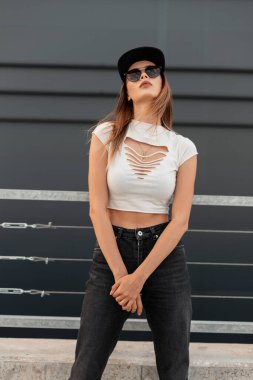 Beyaz stil giyinmiş çekici şehir kadını şık güneş gözlüklü kot pantolonlu ve açık havada gri arka planda modaya uygun siyah şapkalı. Gündelik gençlik kıyafeti..