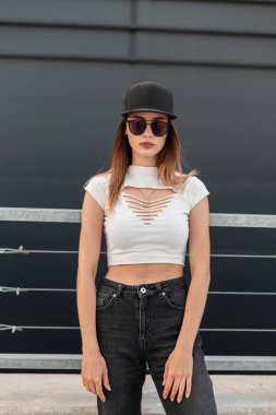 Sıska, seksi, siyah gözlüklü, modaya uygun güneş gözlüklü, yeni koleksiyonundan yaz kıyafetleri ve şehrin klasik duvarının yanında duran siyah beysbol şapkalı genç bir hippi kız..