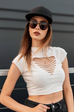 Portre modası güzel şehir kadını şık giyinmiş havalı güneş gözlüklü kot pantolonlu ve açık havada gri arka planda modaya uygun siyah şapkalı. Gündelik gençlik kıyafeti..