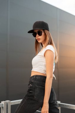 Modern şehirli genç bayan şık siyah Amerikan beysbol şapkalı, modayla seksi, koyu güneş gözlüklü ve klasik kot pantolonlu turuncu yaz günbatımının arka planındaki karanlık binanın yakınlarında poz veriyor.. 