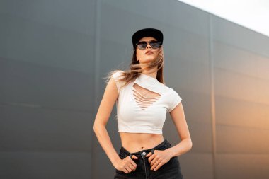 Moda modasına uygun, koyu güneş gözlüklü, şık, havalı genç ve siyah şapkalı seksi manken kadın turuncu gün batımında şehrin klasik duvarının yanında poz veriyor. Sıradan yaz tarzı..