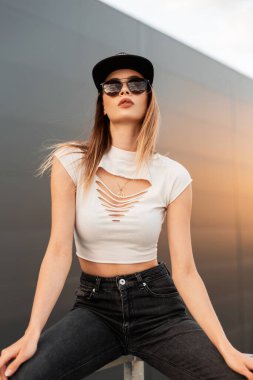 Sıska seksi hippi kız, modaya uygun koyu güneş gözlükleriyle, şık gençlik yaz kıyafetleriyle, yeni koleksiyonundan ve siyah şapkalı, şehrin klasik duvarının yanında oturuyor. Günbatımında havalı kız mankeni.