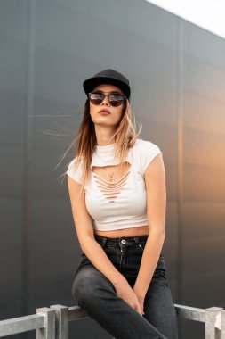 Moda mankeni, güneş gözlüklü genç bir kadın. Yeni koleksiyondaki son moda gençlik kıyafetleri ve siyah şapkalı. Şehrin arka planındaki metal korkuluklarda oturuyor. Hippi kız gün batımında dinleniyor..