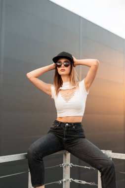 Son moda havalı havalı hippi kız şık şapkalı ve güneş gözlüklü şık genç kız şehirdeki karanlık binanın arka planında metal korkuluklarda oturuyor. Turuncu günbatımında güzel şehir kadını.