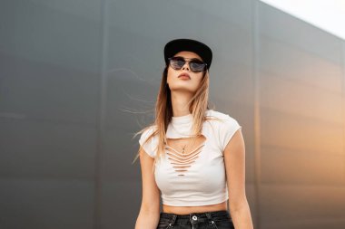 Moda portresi güzel şehir kadını seksi dudakları beyaz stil giyinmiş şık güneş gözlüklü kot pantolonlu ve açık havada gri arka planda siyah beyzbol şapkalı. Yaz gençliği.