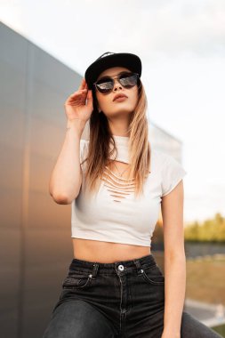 Modayı takip eden modaya uygun siyah şapkalı seksi hippi kız günbatımında modern binanın yakınında şık güneş gözlüklerini düzeltiyor. Gençlik Amerikan tarzı şehircilik.