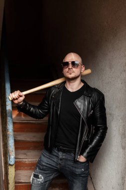 Kotlu siyah deri ceketli, moda gözlüklü, kel bir adam içeride beyzbol sopasıyla ayakta duruyor. Klasik merdivenlerin yanındaki yakışıklı kabadayı..