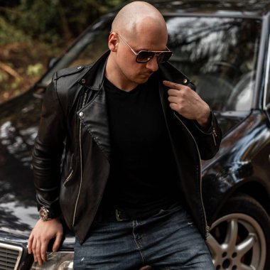 Koyu renk güneş gözlüklü, siyah deri ceketli, modern siyah arabanın önünde şık kotlu havalı bir adam. Güzel bir otomobilin yanında vahşi kel bir adam.