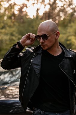 Şık giyinmiş yakışıklı, kel adam gün batımında siyah arabanın yanında duruyor ve güneş gözlüklerini düzeltiyor. Havalı adam şehir dışında ormanda dinleniyor..