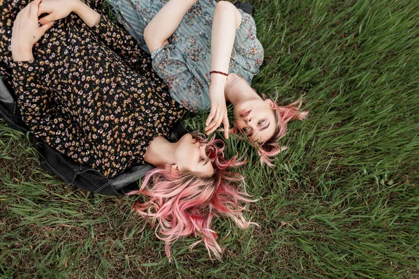新鮮な緑の芝生の背景に花のプリントとスタイリッシュな黒と青のドレスで色の髪を持つ2人の美しい若い女性の上のビュー 可愛いですピンク髪の双子の姉妹は自然の上で芝生の上にあります — ストック写真