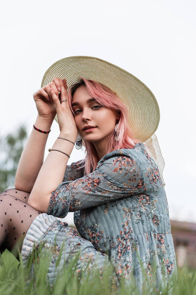Young lovely woman with pink hair in vintage straw hat in stylish summer blue dress sits on fresh green grass outdoors on nature. Beautiful girl fashion model in elegant clothes rests outside city.