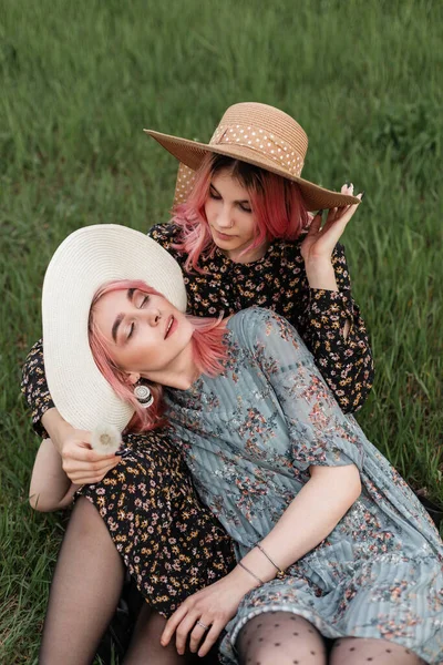 两个穿着时髦青春服装 头戴老式草帽的漂亮女孩躺在城外的草地上 穿着时尚夏装的迷人的辣妹们享受周末的自然之旅 — 图库照片