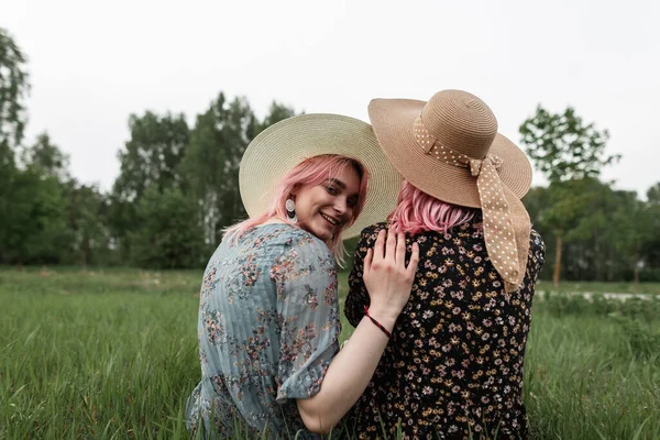 ブルーのドレスでヴィンテージの帽子でかわいい笑顔でピンクの髪を持つモデルの美しい若い女性は妹と座って振り返ります ファッション若者のスタイリッシュなガールフレンドは 夏の日に農村部の芝生の上でリラックスして着用 — ストック写真