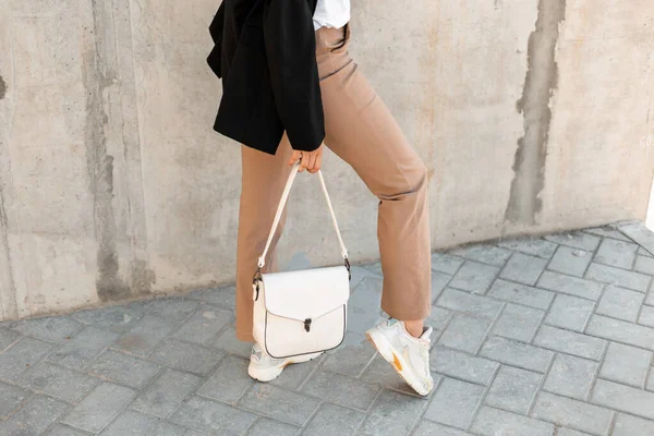 通りの灰色の壁の近くにファッショナブルな白いバッグと白いスタイリッシュなスニーカーを持つ女性 女性のハンドバッグコレクション — ストック写真