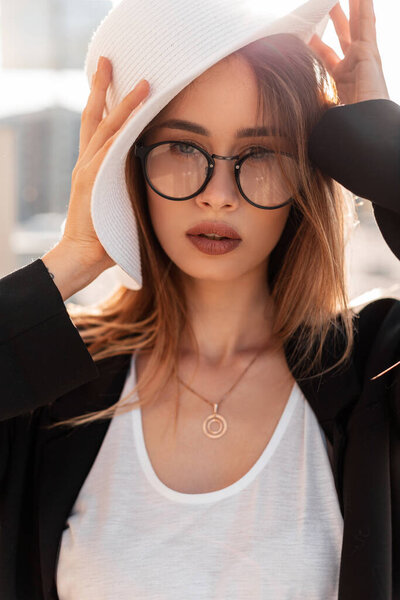 Stylish urban portrait of a young beautiful girl with glasses in a fashionable black blazer and a white T-shirt with a hat in the city at sunset looking at the camera