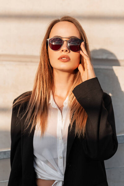 Attractive young woman in black stylish blazer straightens sunglasses and enjoys bright sunshine. Beautiful pretty urban girl in casual vintage clothes posing near building in city at sunset.