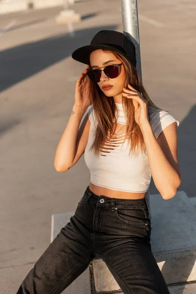 スタイリッシュな白のセクシーな唇を持つファッションの肖像都市の女性は 涼しい流行のサングラスでジーンズでトップを引き裂き 黒い野球帽は日没時に路上駐車場に座っています スワッグ ガール — ストック写真