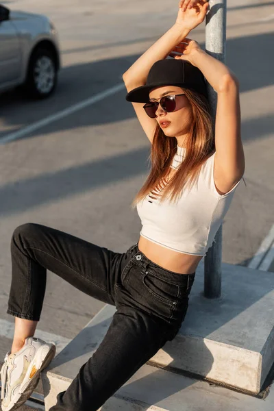 現代的なスタイリッシュな女性で黒のファッショナブルなキャップでTシャツでジーンズでファッションサングラスは晴れた日に路上駐車場で金属柱の近くにリラックス — ストック写真