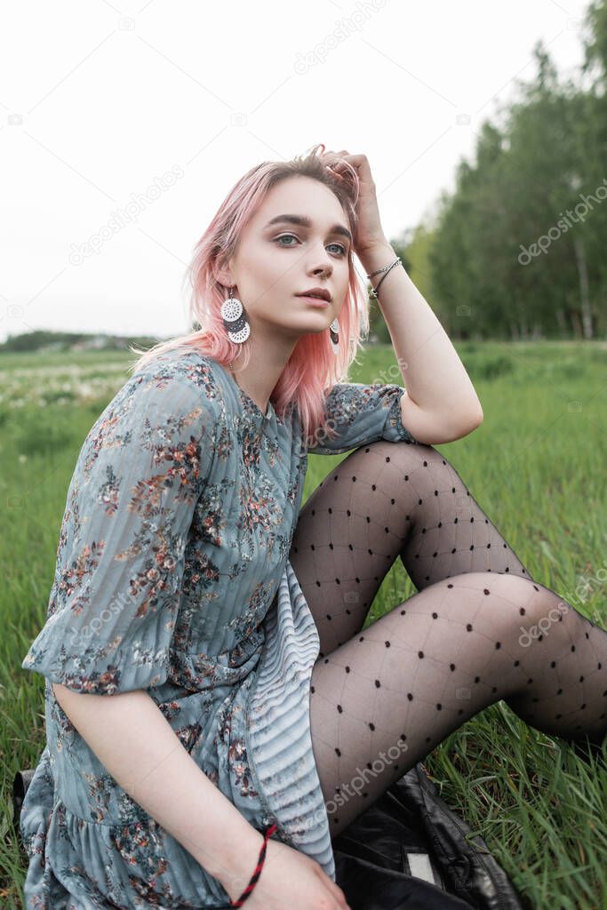 Pretty young woman with pink hair in stylish blue dress in black vintage tights sits on fresh green grass outdoors on nature. Attractive girl fashion model in elegant clothes rests outside city.