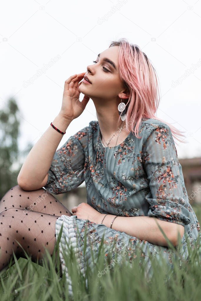 Gorgeous tender young woman with pink hair in fashion elegant blue dress with floral print sit on green grass in field outdoors. Lovely beautiful girl in summer stylish outfit relaxes outside city.