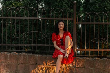 Kırmızı elbiseli güzel bir kadın model şehirde bir çitin yanında oturuyor.